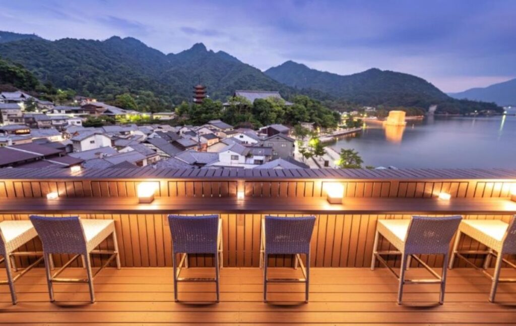 Rooftop terrace bar with wooden counter, rattan stools, overlooking Miyajima waterfront and mountains at dusk