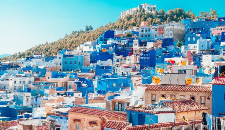 Chefchaouen's iconic blue-painted buildings densely packed on a hillside, with terracotta rooftops and satellite dishes, against a bright turquoise sky.