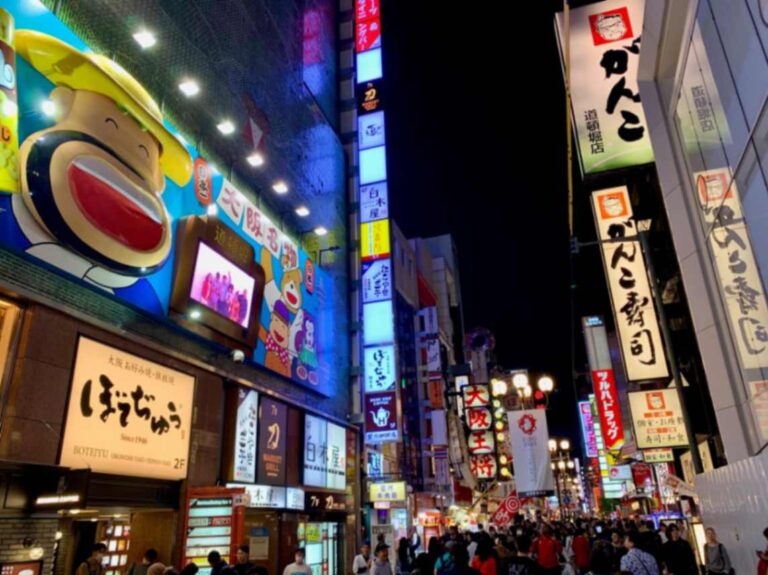 Vibrant night scene of Dotonbori, a famous entertainment district in Osaka, Japan. The street is lined with brightly lit billboards, neon signs, and advertisements in Japanese and English covering multi-story buildings. A large cartoon character with a wide smile dominates one facade. The bustling street is filled with pedestrians, capturing the lively atmosphere of Osaka's iconic nightlife and food scene