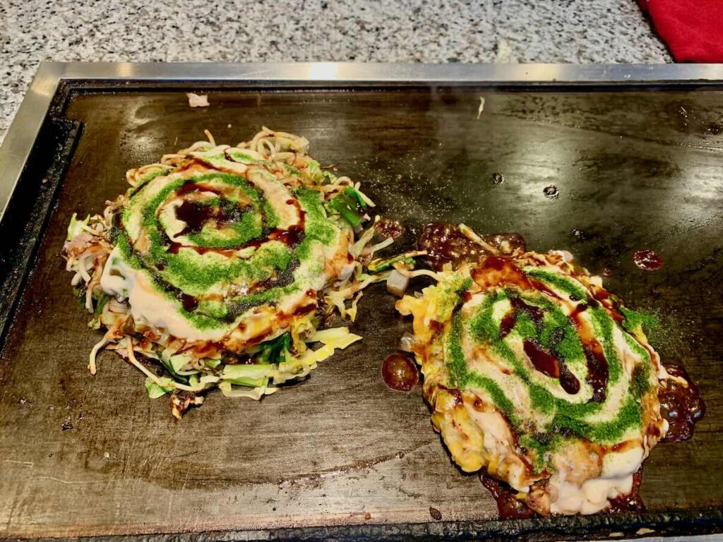 Two okonomiyaki cooking on the teppan at Mizuno in Osaka, topped with brown sauce, mayonnaise and green aonori seaweed