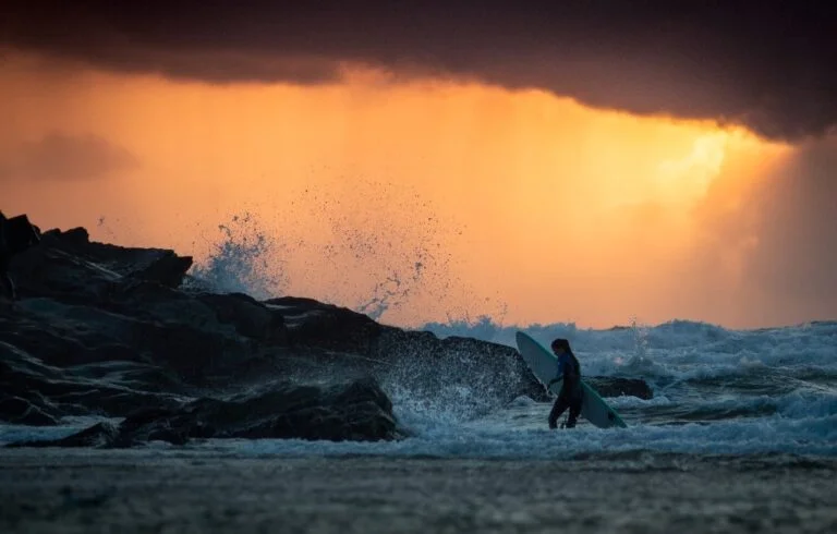 Surfer walking in the waves crashing off rocks in the sunset light. Polzeath Beach Cornwall