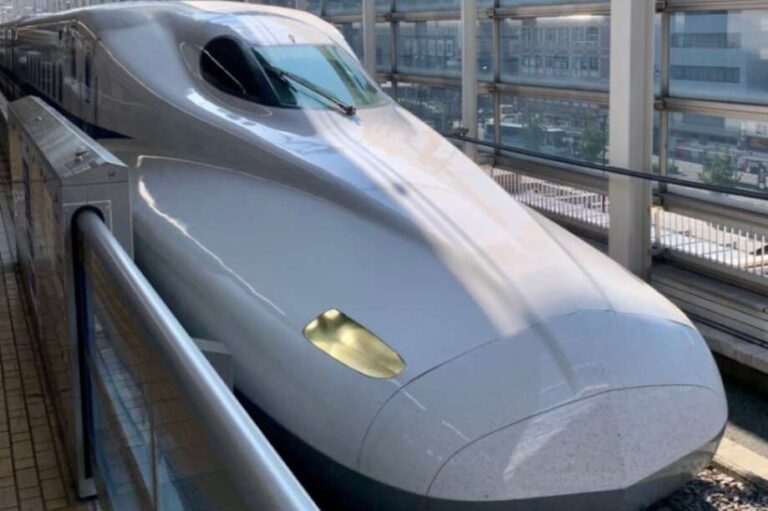 Close-up view of a sleek, white Japanese Shinkansen bullet train at a station platform. The train's aerodynamic nose cone is prominently featured, showcasing its streamlined design. The image captures the train's modern, high-speed appearance, with a glimpse of the station and city buildings visible through the platform's glass enclosure in the background