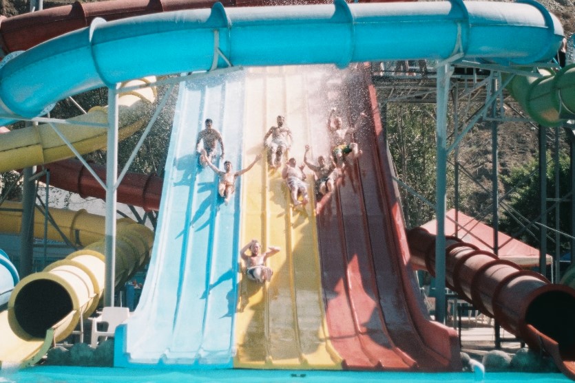 Children and adults alike enjoy a thrilling multi-lane water slide at a vibrant waterpark. The slide is brightly colored in blue, yellow, and red lanes, surrounded by twisting enclosed tube slides. The setting is lively, with splashing water and clear blue skies, creating a fun and adventurous atmosphere