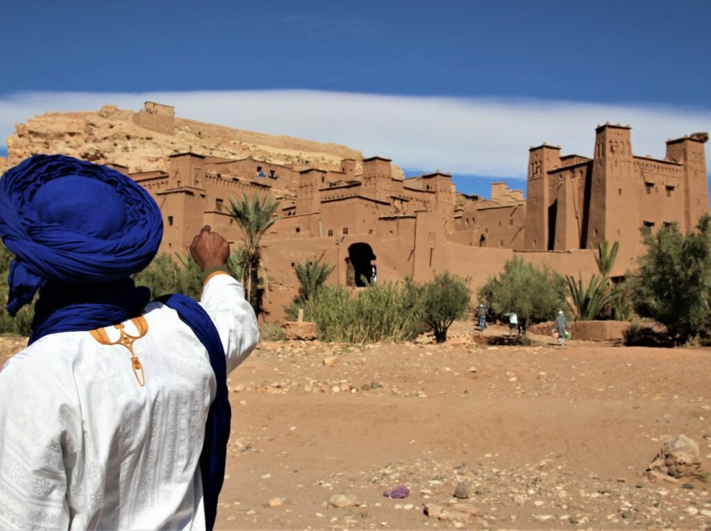 Berber guide in white robe and blue turban points up towards the earthen-clay towers of Ait Benhaddou kasbah in Morocco