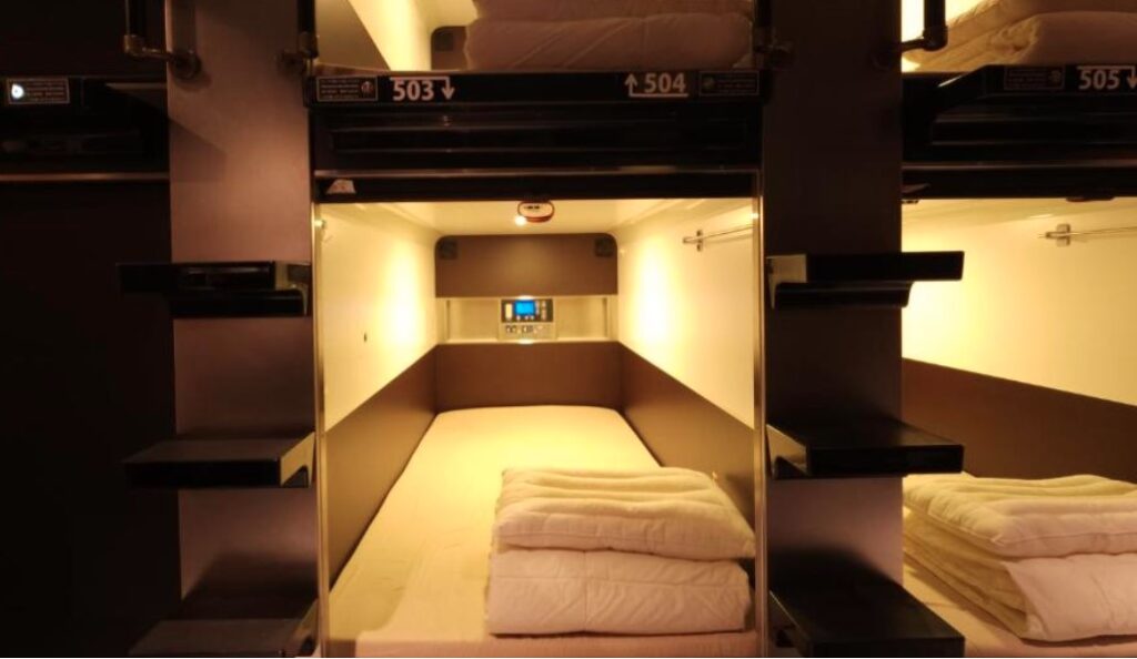 Capsule-style dormitory bed pod with warm lighting, white bedding, and numbered compartments