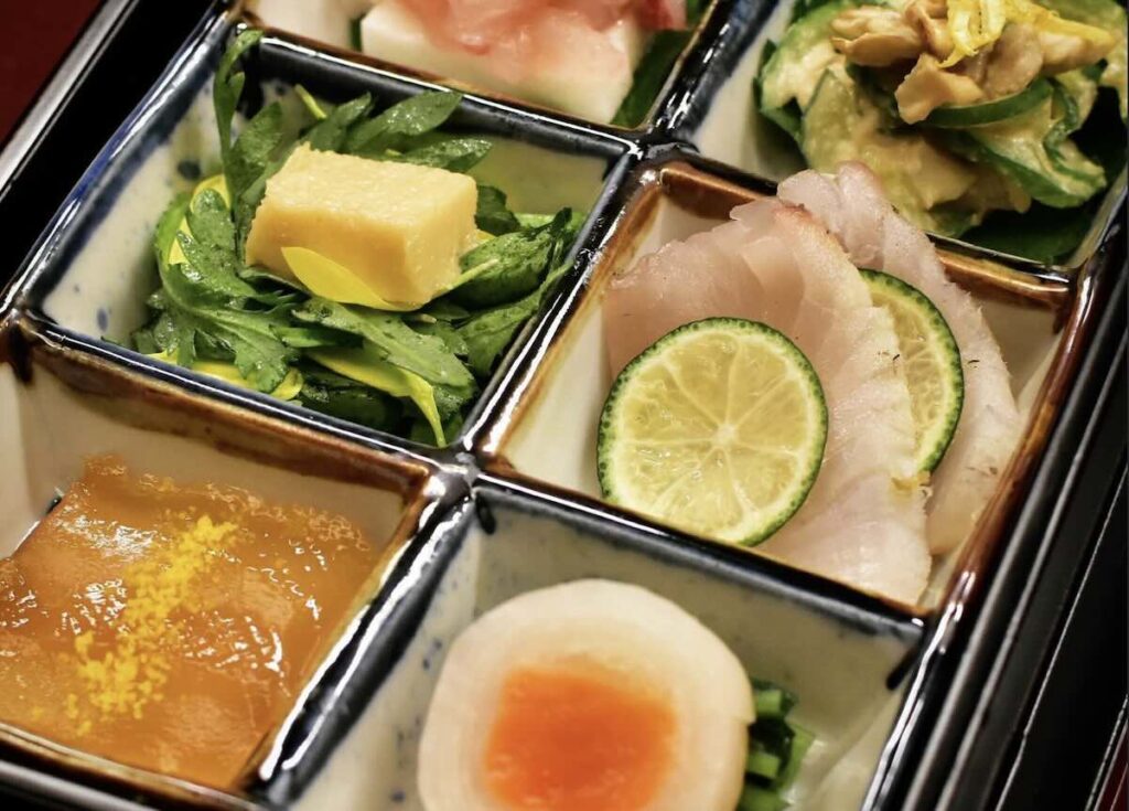 A compartmentalised ceramic tray holding kaiseki appetisers: sashimi with sudachi, tamagoyaki, simmered egg, and seasonal vegetables. 