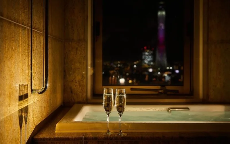 A luxurious hotel bathroom with a bathtub, featuring two champagne flutes in the foreground and a nighttime view of the Tokyo Skytree through the window.