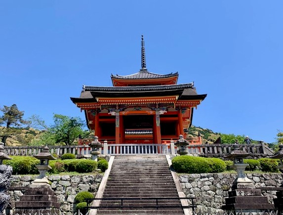 A grand Japanese temple entrance with a vibrant red and orange multi-tiered roof. Stone steps lead up to the ornate structure, flanked by stone lanterns and meticulously trimmed shrubs. The temple is set against a backdrop of rocky hills and clear blue sky.