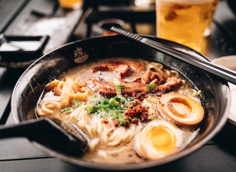 A close-up of a bowl of ramen, featuring a rich, creamy broth, tender slices of pork, soft-boiled eggs, green onions, and a sprinkle of chili flakes. The noodles are thick and inviting, with chopsticks resting on the bowl’s edge. In the background, a blurry image of a drink can be seen, adding to the cozy dining atmosphere.