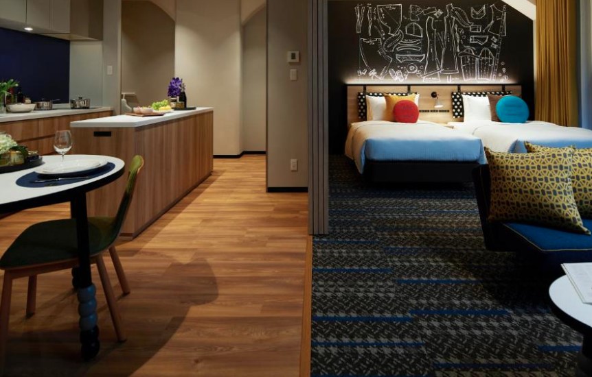Modern hotel suite with kitchenette and dining area connected to a twin bedroom featuring abstract wall art, colourful cushions and carpeting