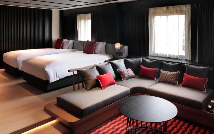 Spacious twin room with black panelled walls, large L-shaped sofa with red cushions, and black coffee table on red rug