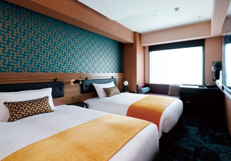 Twin hotel room with turquoise geometric wall feature, orange bed runners and patterned cushions, facing large window