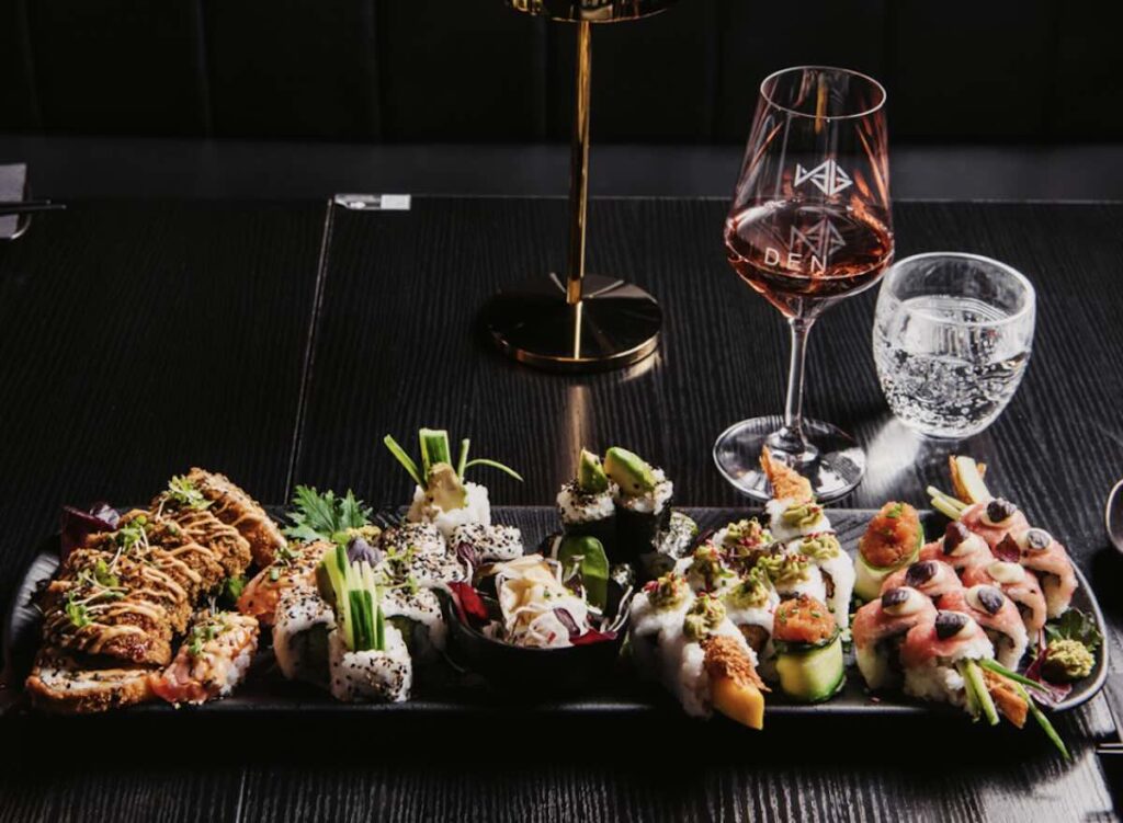 A long platter of assorted sushi rolls with avocado, salmon, and various toppings, alongside a glass of rosé wine on a dark table. 