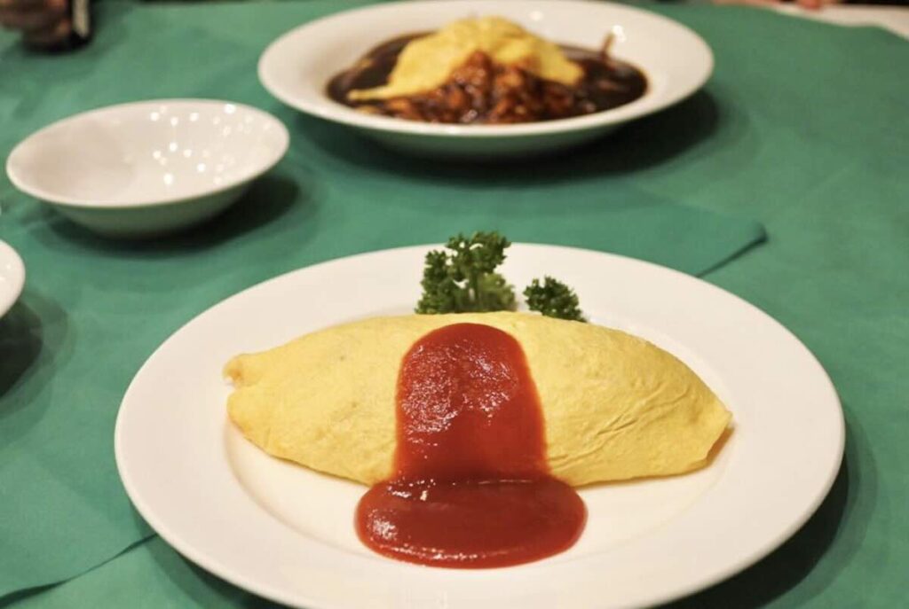 A classic omurice, an omelette wrapped around rice, topped with ketchup and parsley, with a hashed beef dish in the background. 