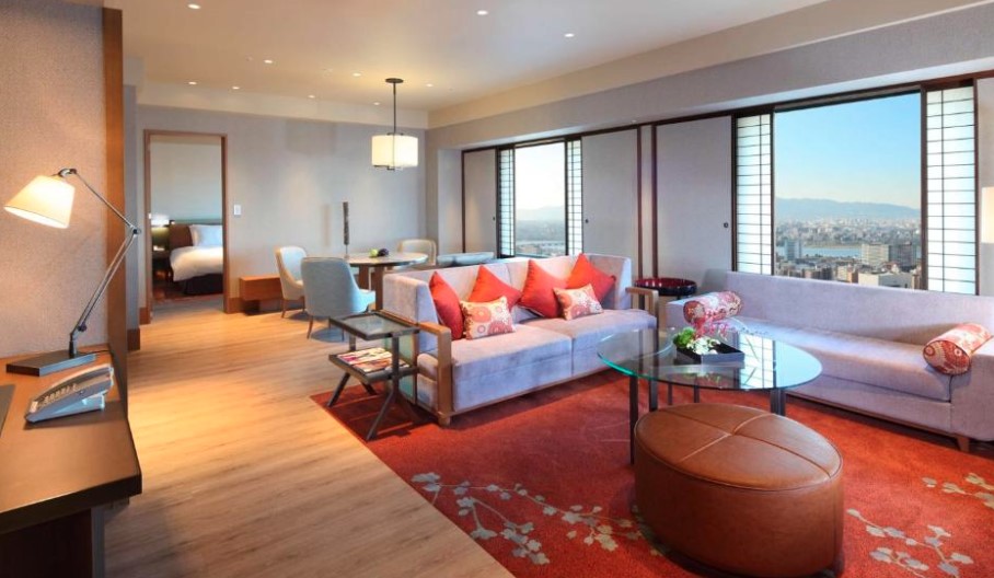 Luxury suite with city views through floor-to-ceiling windows, featuring grey sofas with red cushions and a desk area
