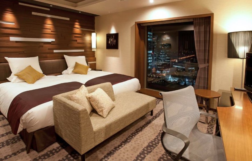 Twin room with textured wooden headboard wall, night cityscape view, beige sofa and brown bed runners