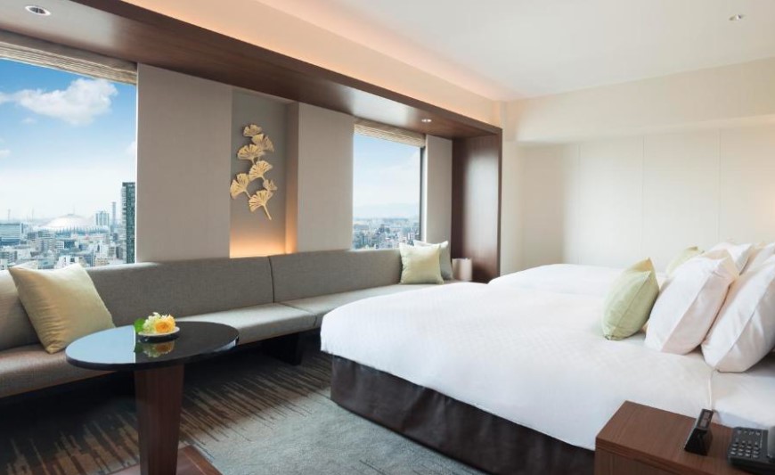 Modern hotel room featuring king bed, built-in grey sofa, and metallic ginkgo leaf wall art. Floor-to-ceiling windows showcase city skyline
