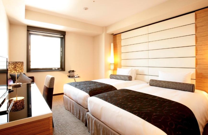Modern twin room with wooden furnishings, black and white bedding, geometric headboard panel and desk area near window.