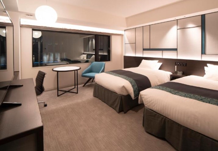 Modern twin room with geometric light panel, blue accent chair and desk area. Features round table and spherical pendant light