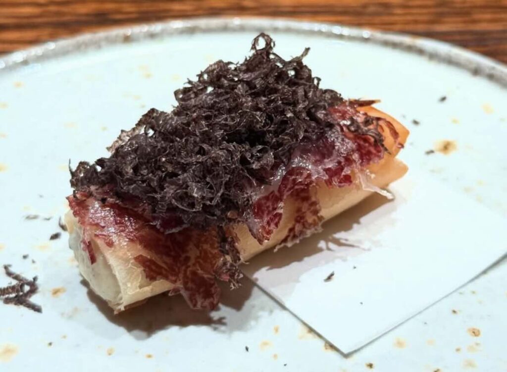 A piece of nigiri sushi topped with marbled wagyu beef and a generous mound of freshly shaved black truffle, on a pale ceramic plate. 