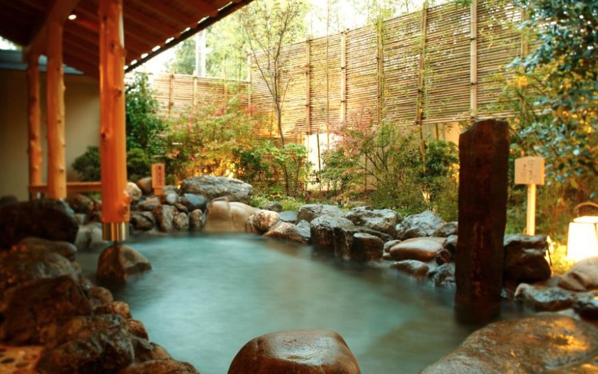 Outdoor onsen bath surrounded by natural rocks and bamboo fencing. Steam rising from mineral water, lantern lighting.