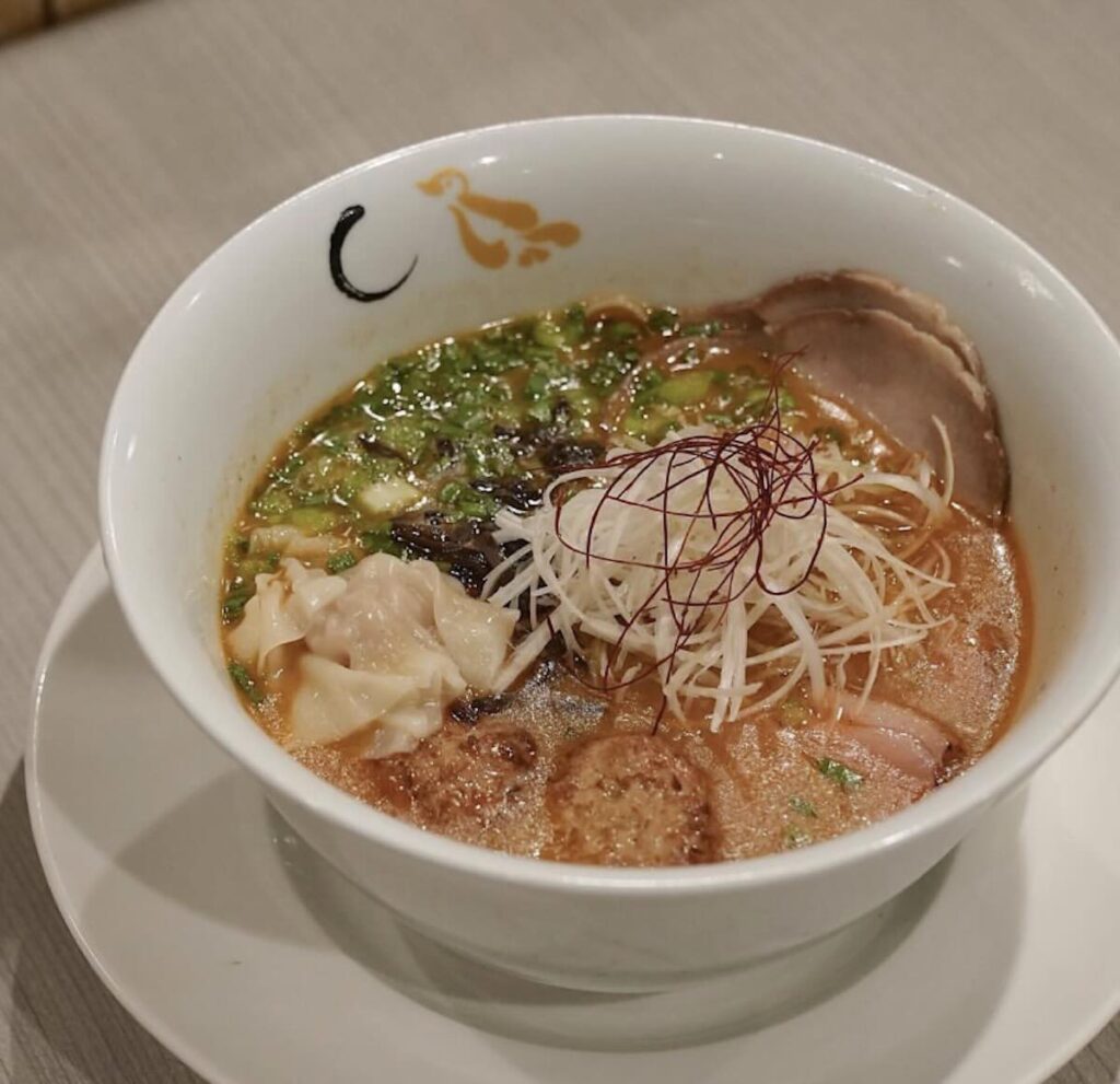 A bowl of rich ramen topped with chashu, a wonton, shredded white leek, red chilli threads, and spring onions, on a saucer. 