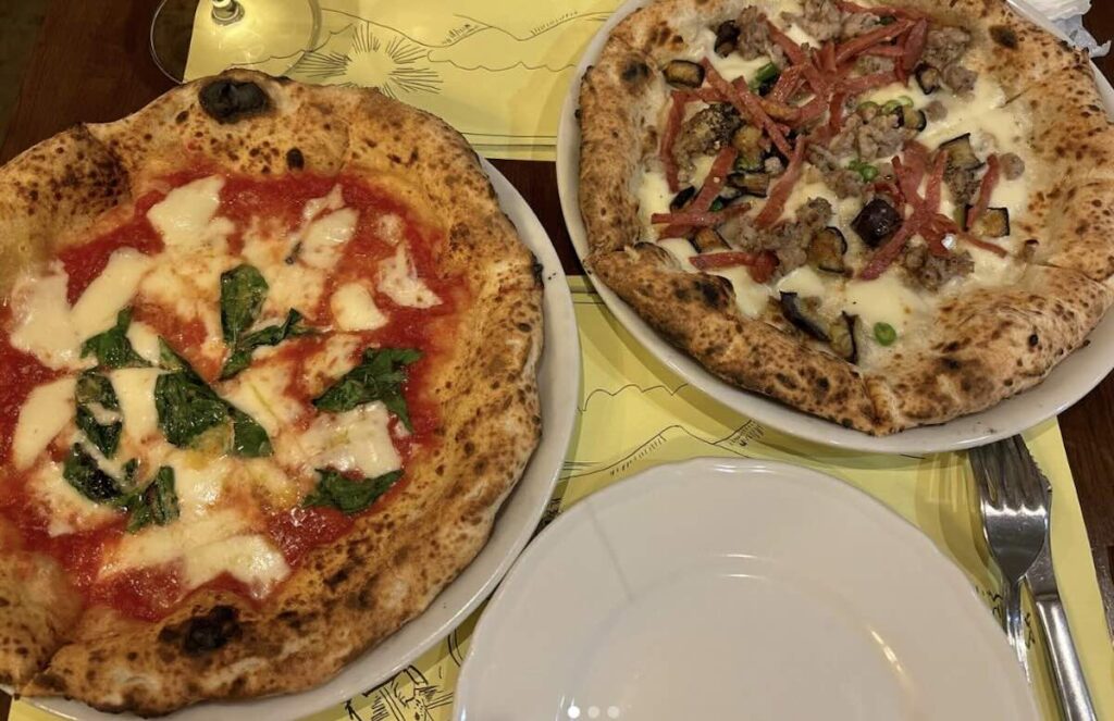 Two Neapolitan pizzas on a yellow placemat: a margherita with basil, and a white pizza with aubergine, sausage, and salami. 