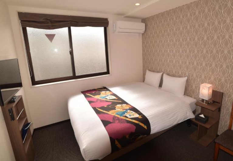Compact hotel room with double bed featuring colourful Japanese-style bed runner, wall-mounted TV and patterned wallpaper