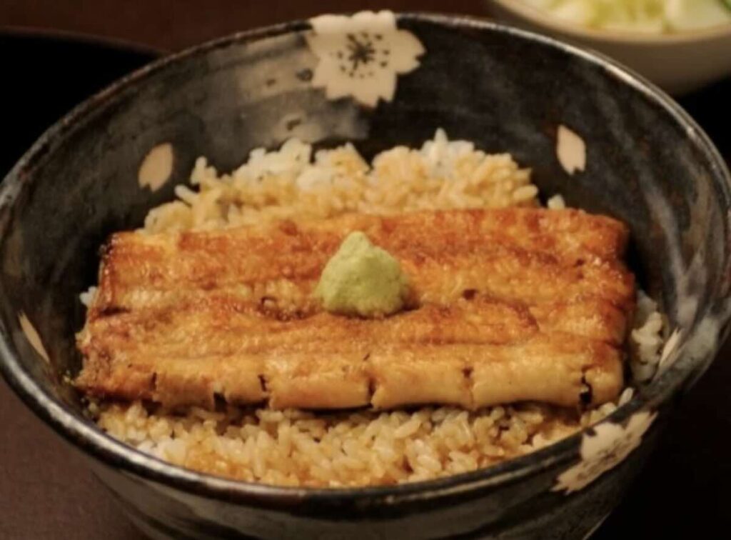 A bowl of grilled eel over rice topped with a small mound of fresh wasabi, served in a dark ceramic bowl. 