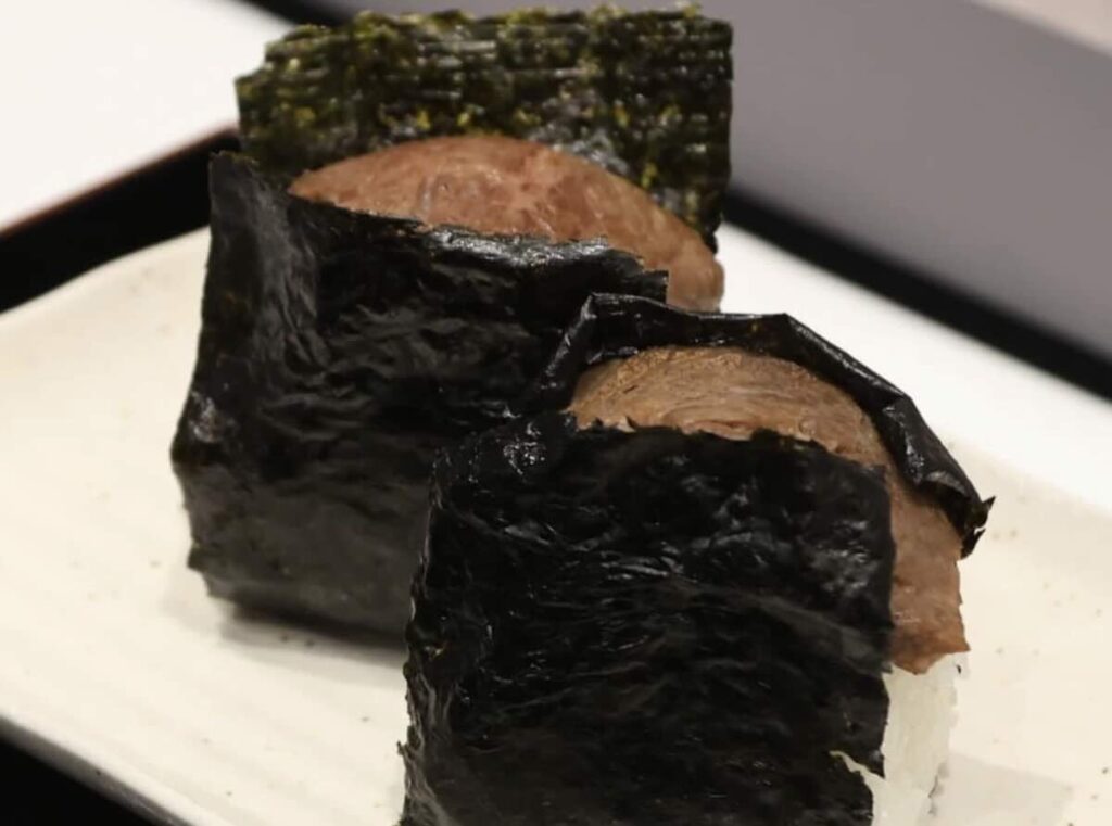 Two onigiri wrapped in dark nori, each topped with a slice of slow-cooked beef, served on a white rectangular plate.