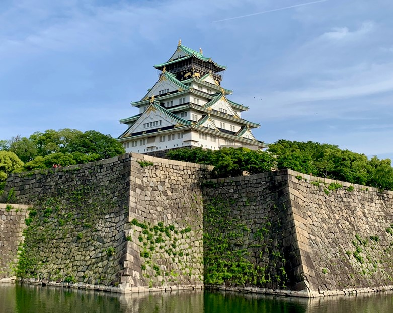Majestic Osaka Castle rising above its stone walls and moat. The iconic Japanese fortress features a white and green multi-tiered pagoda roof atop a stone base, surrounded by lush greenery and reflected in calm waters below on a clear, sunny day.