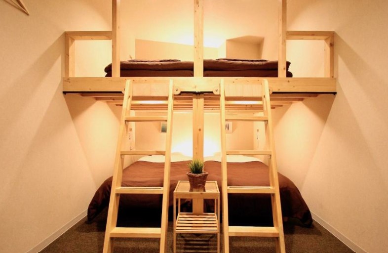 Compact hostel-style room with wooden bunk beds, brown bedding and central ladder access. Small plant on side table between beds.
