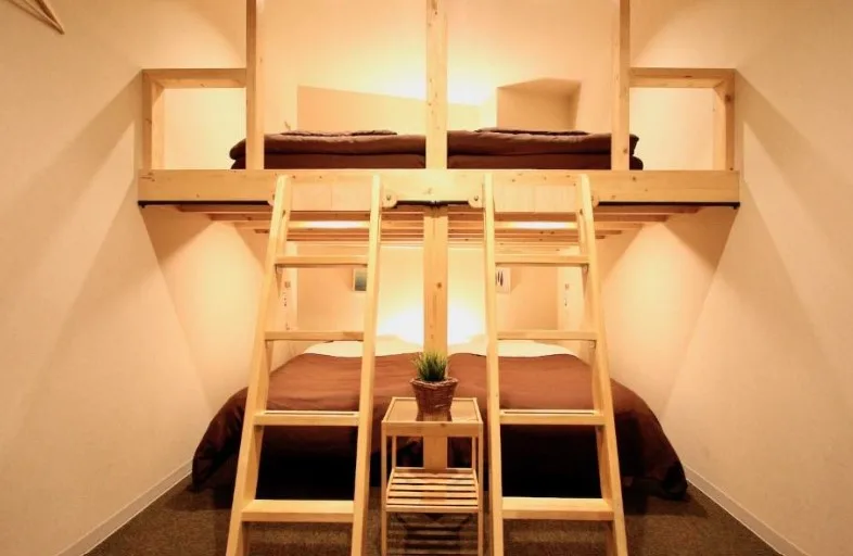 Compact hostel-style room with wooden bunk beds, brown bedding and central ladder access. Small plant on side table between beds.