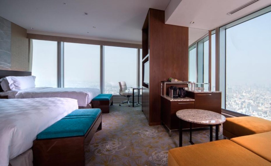 Twin hotel room with floor-to-ceiling windows, white bedding, teal bed runners and orange sofa. Wooden partition with minibar