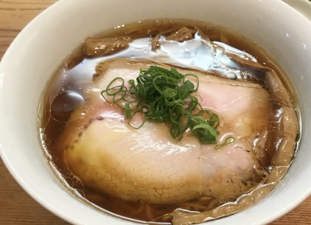 A bowl of clear shoyu ramen with a slice of pink-edged chashu pork, finely sliced spring onions, and bamboo shoots. 