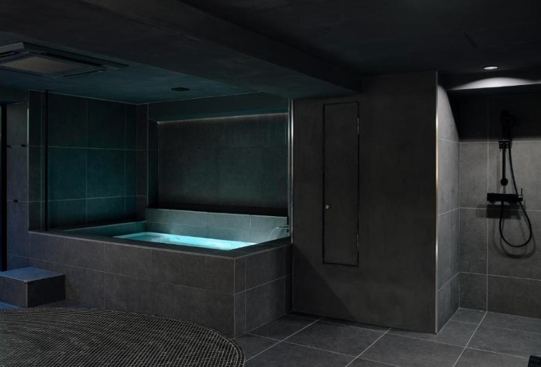 Modern spa room with illuminated plunge bath and shower area. Dark grey tiles throughout, ambient lighting.