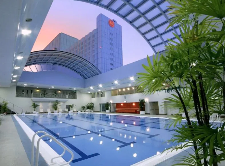 Indoor swimming pool under curved glass roof. Palm plants frame the pool, hotel tower visible through arched ceiling at sunset.
