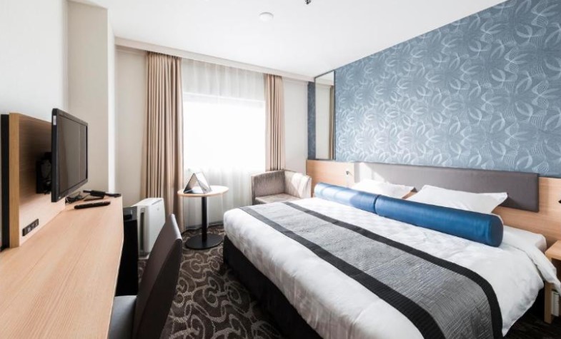 Double room with patterned blue feature wall, wooden desk and grey carpeting. Blue bolster pillow on white bedding