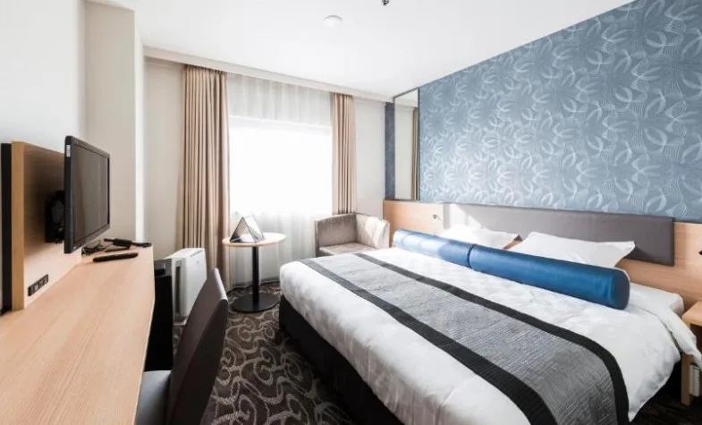 Double room with patterned blue feature wall, wooden desk and grey carpeting. Blue bolster pillow on white bedding