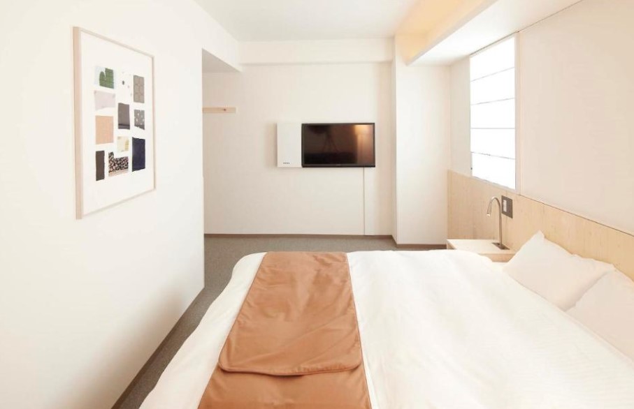 Minimalist double room with tan bed runner, wall-mounted TV and abstract artwork. Features light wood panelling