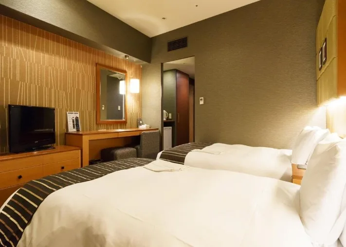 Contemporary hotel room with two white-linened beds, striped bed runners, and wooden headboards. Features textured gold wallpaper, built-in vanity with mirror, flat-screen TV, and olive green walls. The room demonstrates modern business hotel styling