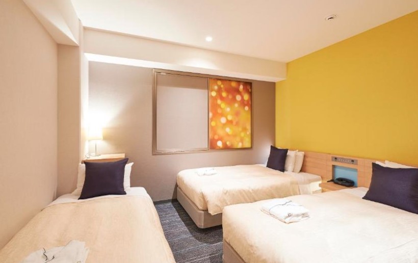 Triple hotel room with yellow accent wall, three single beds with navy cushions, and illuminated artwork panel