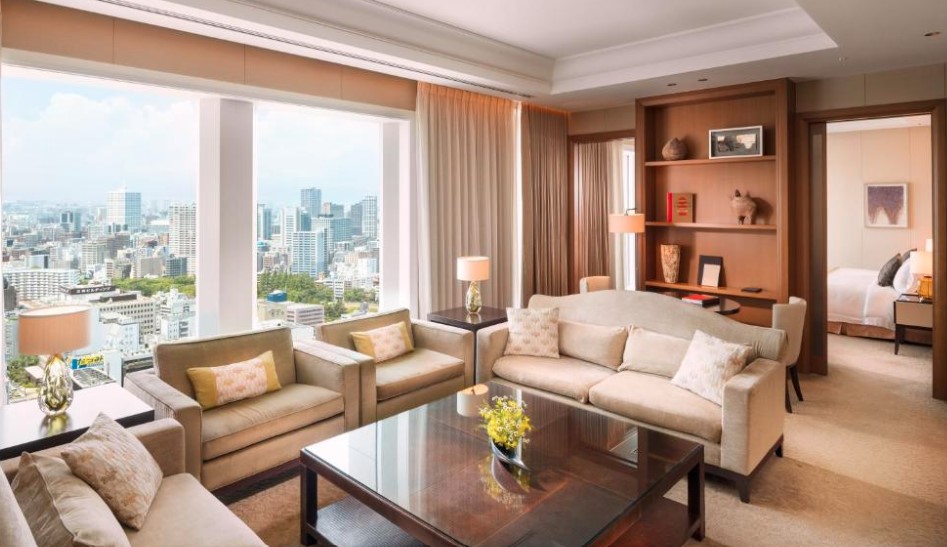 Luxurious hotel suite living room with cream sofas, floor-to-ceiling windows offering panoramic Osaka city views, and built-in wooden shelving.