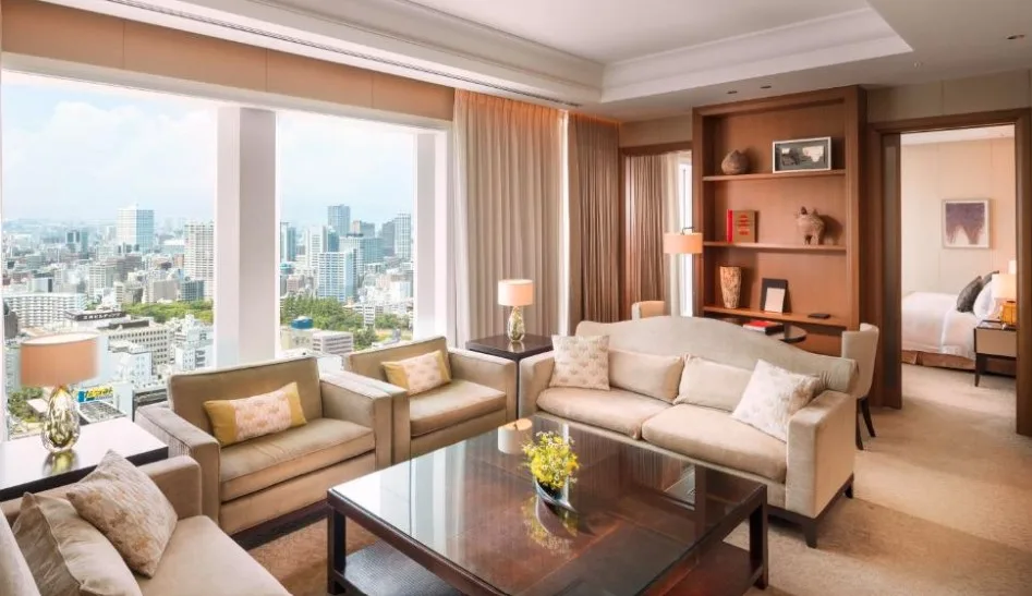 Luxurious hotel suite living room with cream sofas, floor-to-ceiling windows offering panoramic Osaka city views, and built-in wooden shelving.