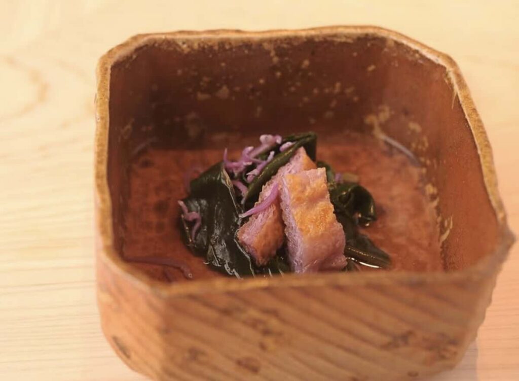 A small rustic ceramic bowl holding wakame seaweed with cubes of seared duck breast and pickled red onion. 