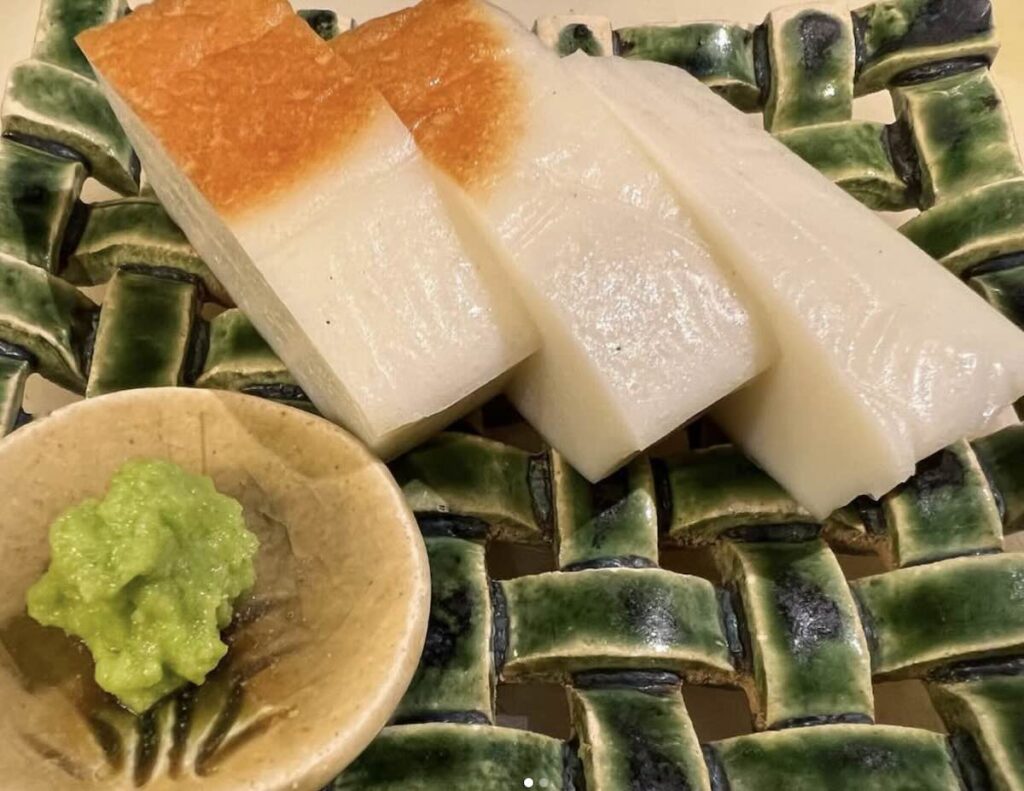 Three thick slices of handmade soba gaki, lightly toasted on top, served on a green woven ceramic plate with fresh wasabi.