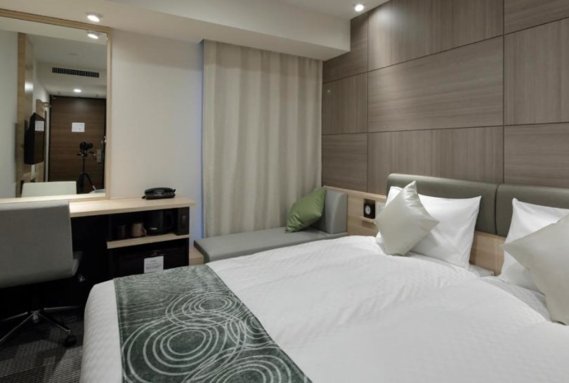 Contemporary hotel room with grey wood panels, white bedding, green accents and built-in desk area. Floor-length curtains