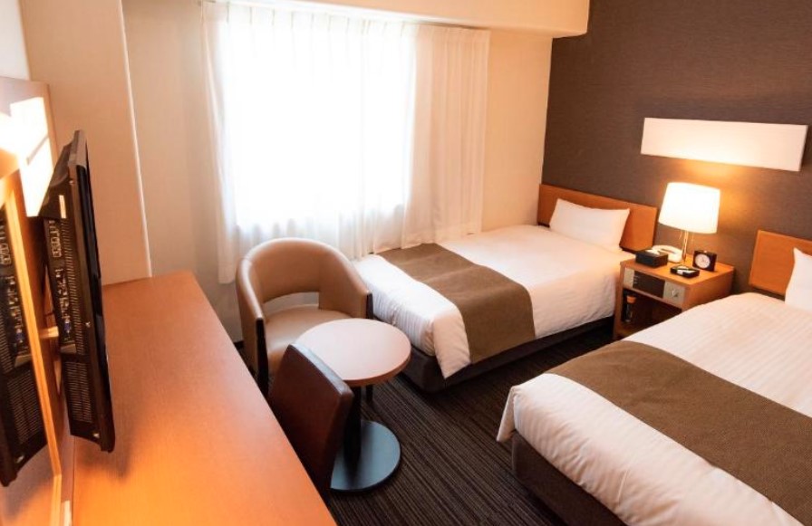 Compact twin room with brown accents, curved armchair and side table. Built-in desk with dark carpeted flooring