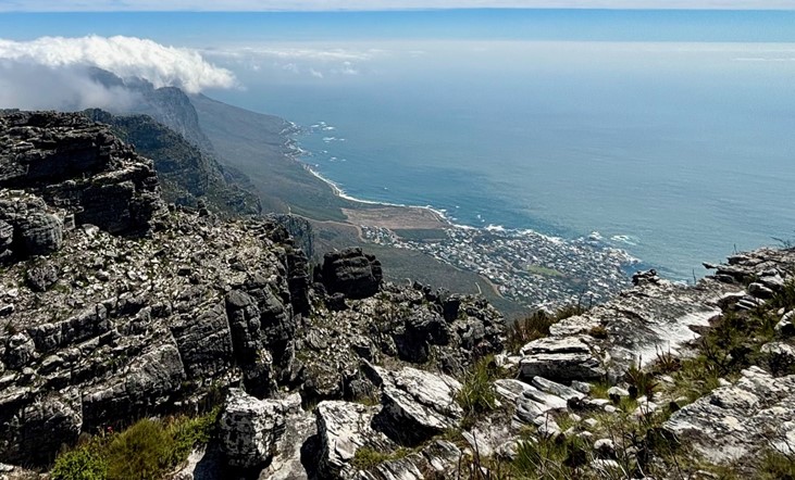 A breath taking view from the rock top of Table Mountain, Cape Town, looking out over the coastline and the ocean in Camps Bay. The sky is vibrant blue with scattered wispy clouds and a layer of fog rolls over the mountains in the distance