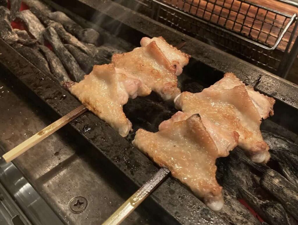 Two skewers of chicken thigh grilling over binchotan charcoal, the skin crisping golden brown above glowing embers. 